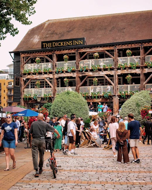 A busy outdoor scene in front of a large, multi-story wooden building called 'The Dickens Inn' with a sign on the roof, featuring exposed timber framing and decorated with flower boxes on each level.